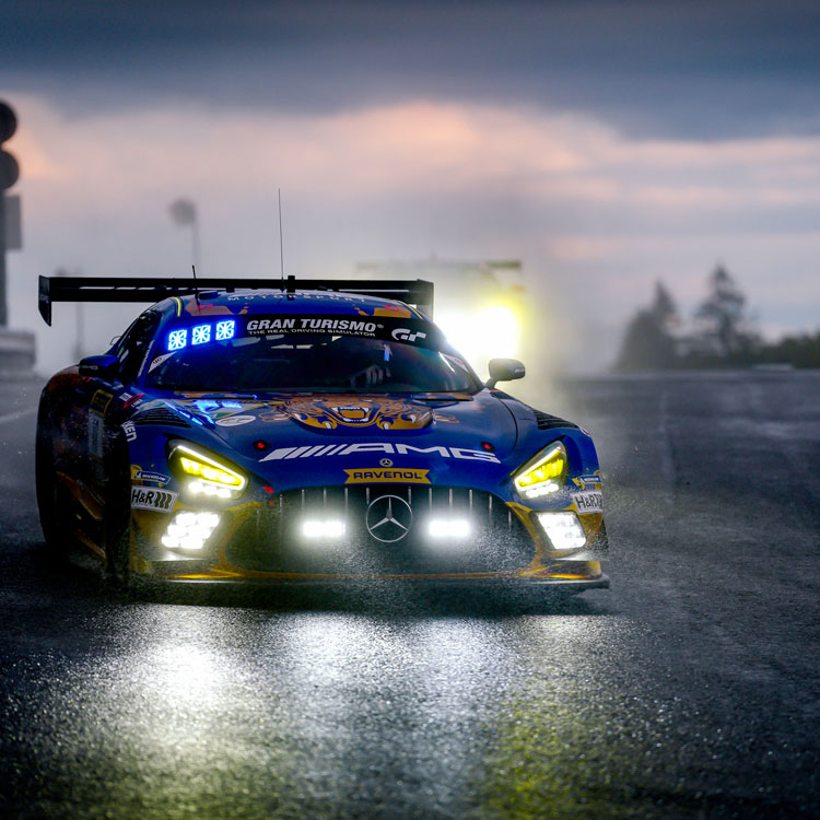 Blue AMG GT racing car with bright headlights on wet track during dark cloudy hours.