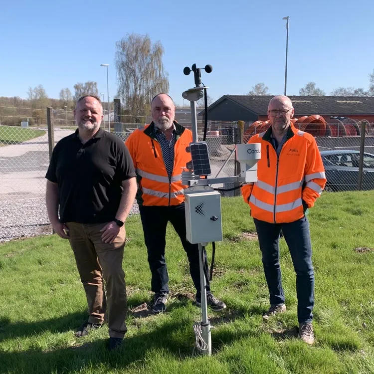 Mads and employees from Arkil standing outdoors on green grass in front of a fenced area with industrial buildings; the two men from Arkil are wearing orange reflective jackets and one wears a black shirt; in front of them is a weather station from cordulus with solar panels and sensors.