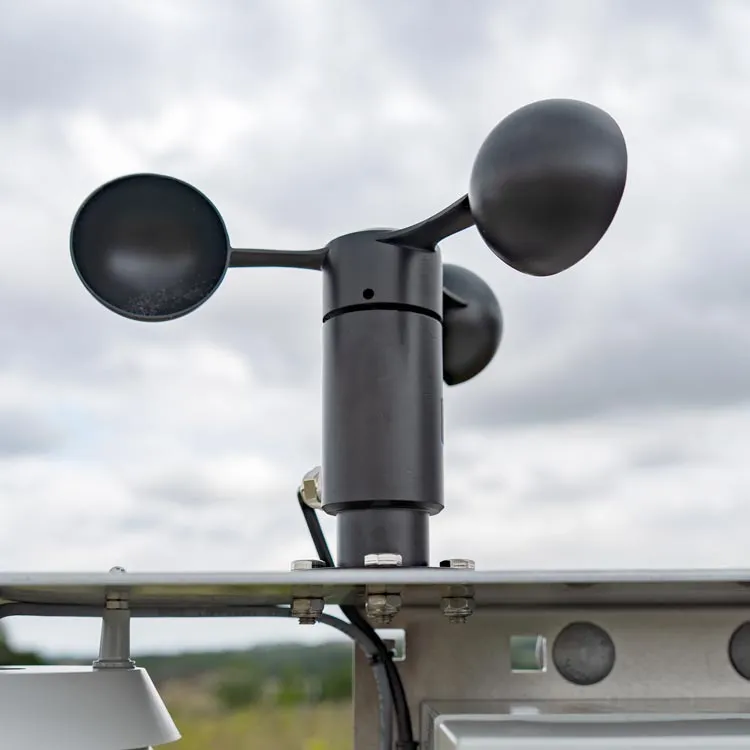 Close-up of a black anemometer from cordulus site station, a device with three cup-shaped arms used to measure wind speed, mounted on a metal structure outdoors under a cloudy sky.