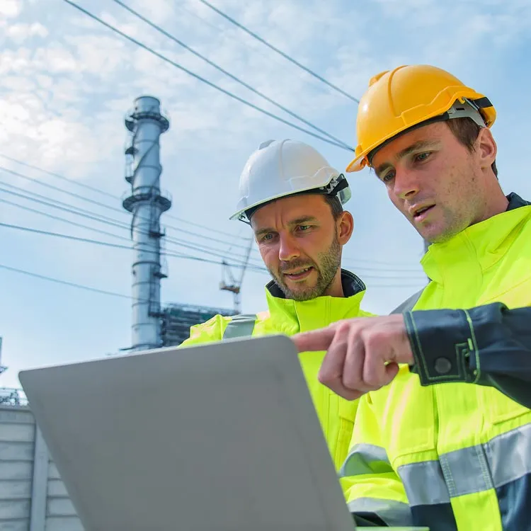 Two engineers in yellow safety jackets and helmets, one white and one yellow, looking at a laptop outdoors with an industrial plant and power lines in the background under a partly cloudy sky.