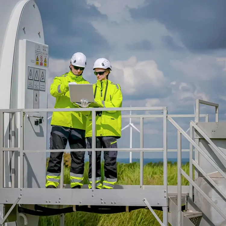 Two workers wearing white helmets, sunglasses, and bright yellow safety jackets standing on a metal platform next to a wind turbine, looking at a laptop under a cloudy sky with another wind turbine in the distance.