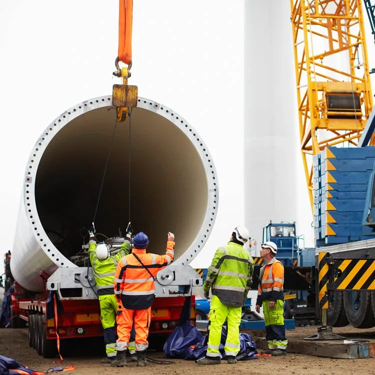 Construction workers in high-visibility gear guiding a large cylindrical structure being lifted by a crane at a worksite.