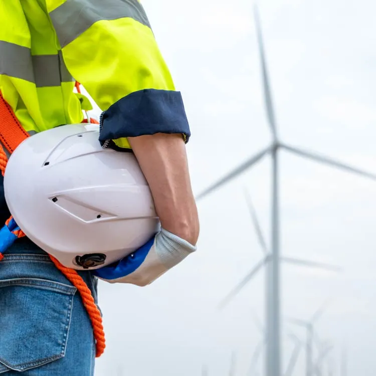 Construction worker wearing a neon yellow jacket and jeans holding a white safety helmet with wind turbines visible in the background under an overcast sky.
