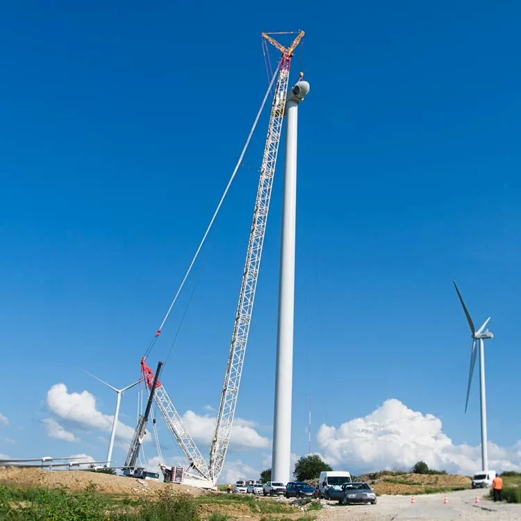 Duży dźwig montujący wieżę turbiny wiatrowej w słoneczny dzień przy czystym, błękitnym niebie; w tle widać inne turbiny wiatrowe i pojazdy.