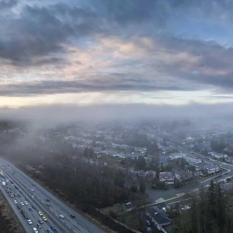A foggy morning aerial view over a suburban neighborhood with a highway on the left filled with cars, under a cloudy sky with patches of light from sunrise or sunset.