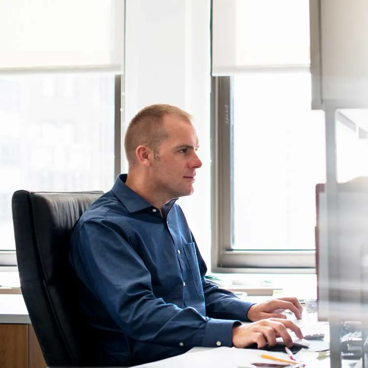 Man in a blue shirt sitting at a desk, using a computer mouse and keyboard, facing a monitor with windows in the background.