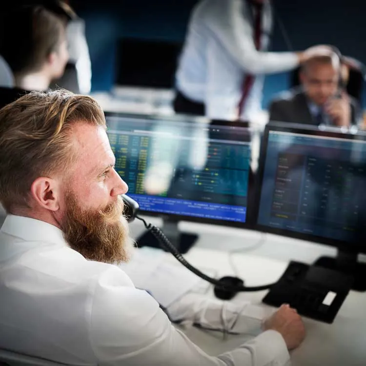 Man with beard and white shirt sitting at a desk using a phone headset, looking at computer monitors displaying data charts, in a busy office environment with colleagues in the background.