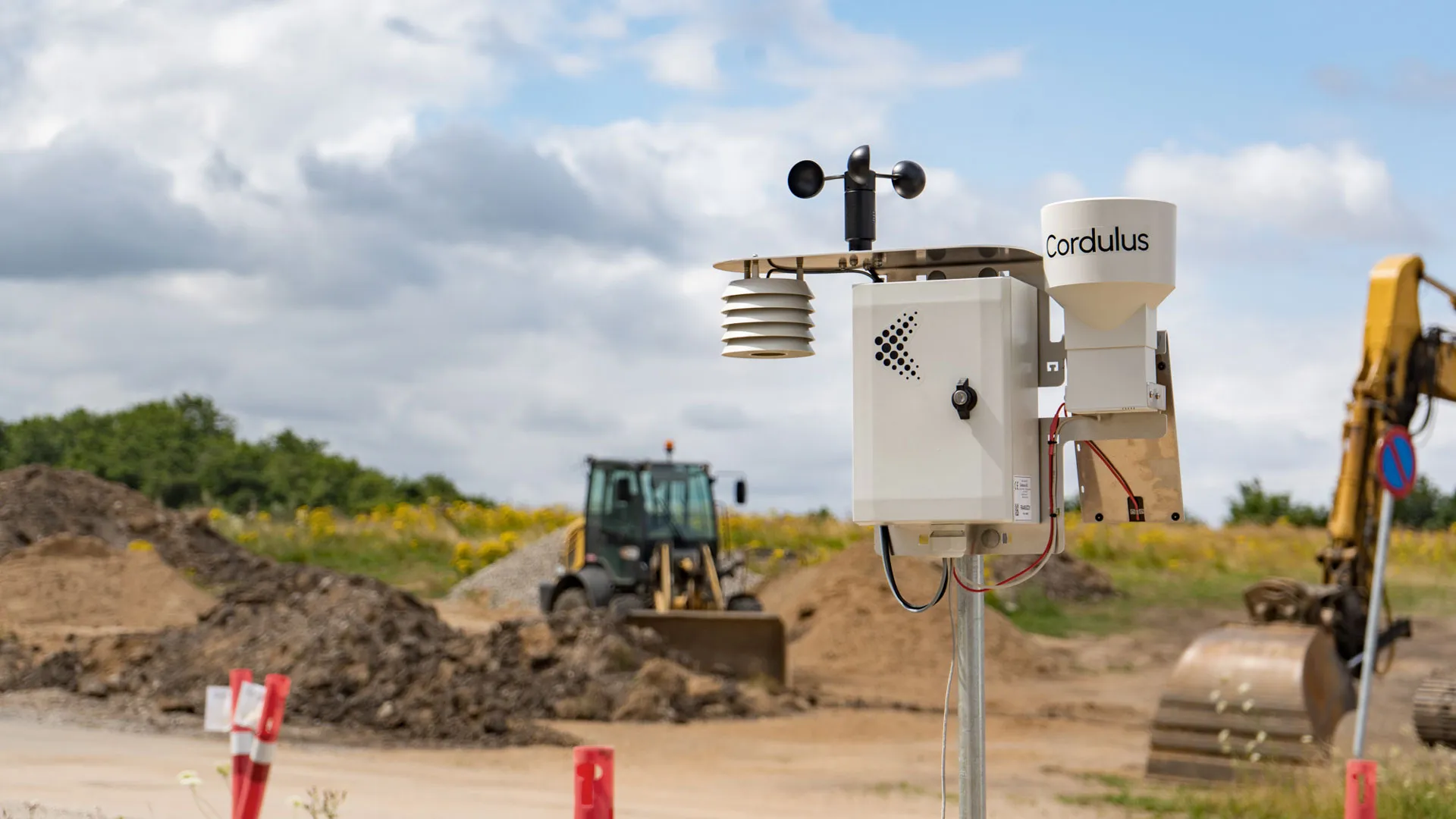 Cordulus Site weather station with instruments including anemometer and radiation shield on a pole at an outdoor construction site with earth-moving equipment and dirt piles in the background under a partly cloudy sky.