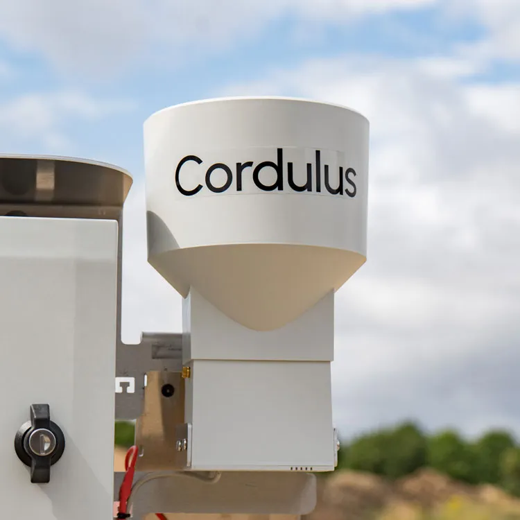 Close-up of a white Cordulus Site rain gauge device mounted outdoors with a partly cloudy sky and greenery in the background.