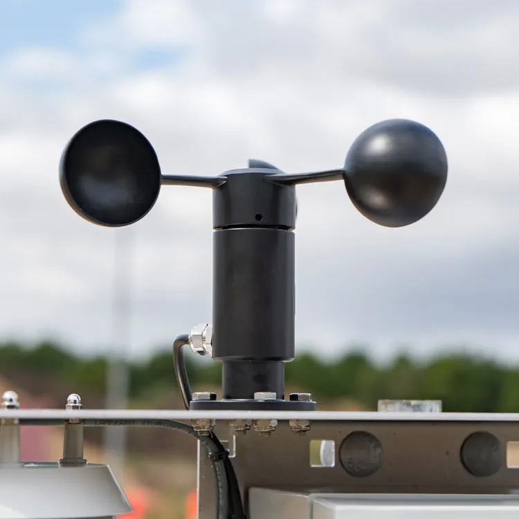 Close-up of a black anemometer mounted on Cordulus Site weather station outdoors, with two cups and a spherical sensor against a cloudy sky background.