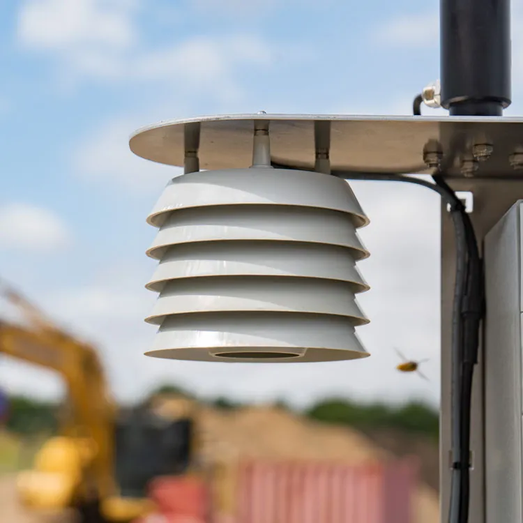 Close-up of a white air sensor with layered shielding mounted on Cordulus Site weather station, with blurry industrial background and a small flying insect nearby.