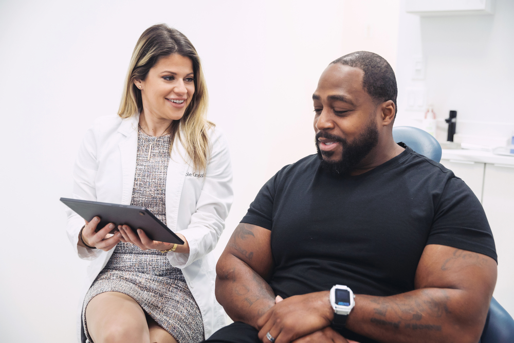 A smiling doctor showing a tablet to a male patient during a medical consultation in a modern clinic, highlighting professional healthcare and patient care services.