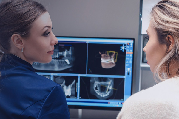 Dentist explaining dental X-ray images to a female patient with tooth and jaw scans on screen.
