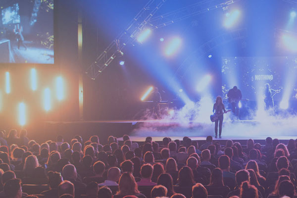 Musicians performing on stage with bright lights and fog in front of a seated audience.