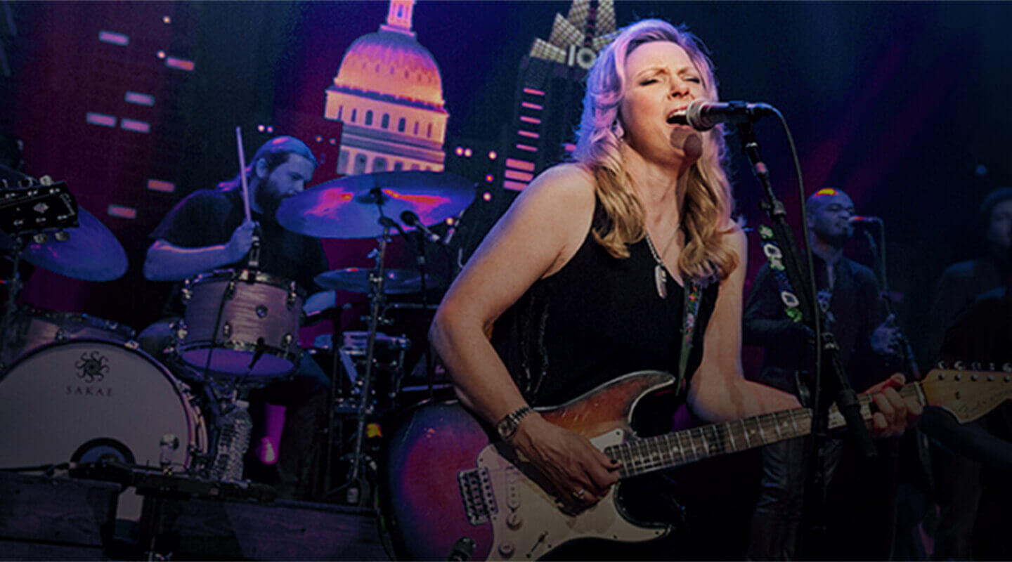 Female musician singing and playing electric guitar on stage with drummer and backup singers behind.