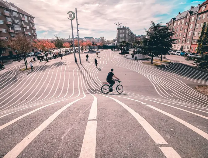 Cyklist krydser en åben plads, hvor svungne hvide striber er malet på asfalten