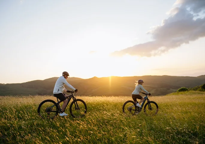 To cyklister på elcykler kører gennem højt græs i et bakket landskab ved solnedgang