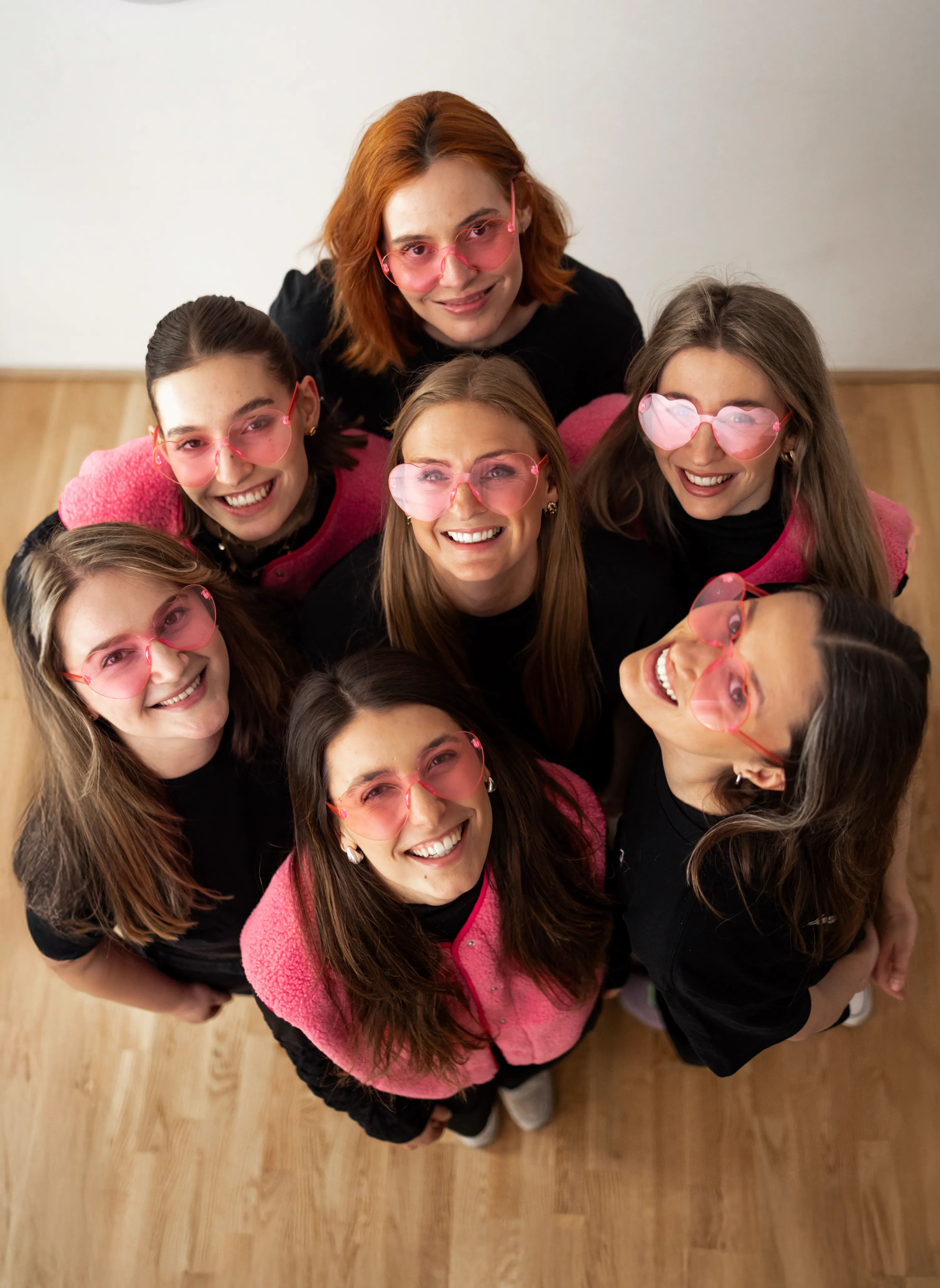 Das A.Quadrat Team von oben fotografiert – sieben lächelnde Frauen mit rosa Herzbrillen