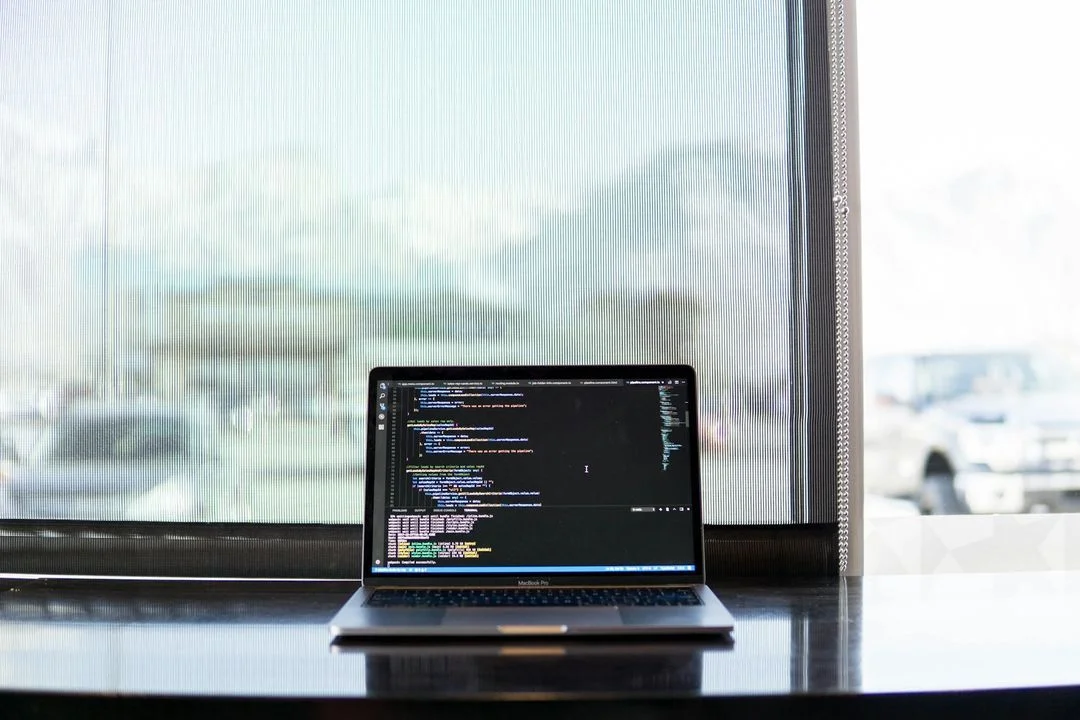 Un ordinateur portable ouvert affichant un éditeur de code sur son écran, posé sur un bureau sombre et réfléchissant devant une fenêtre lumineuse aux stores semi-transparents fermés.