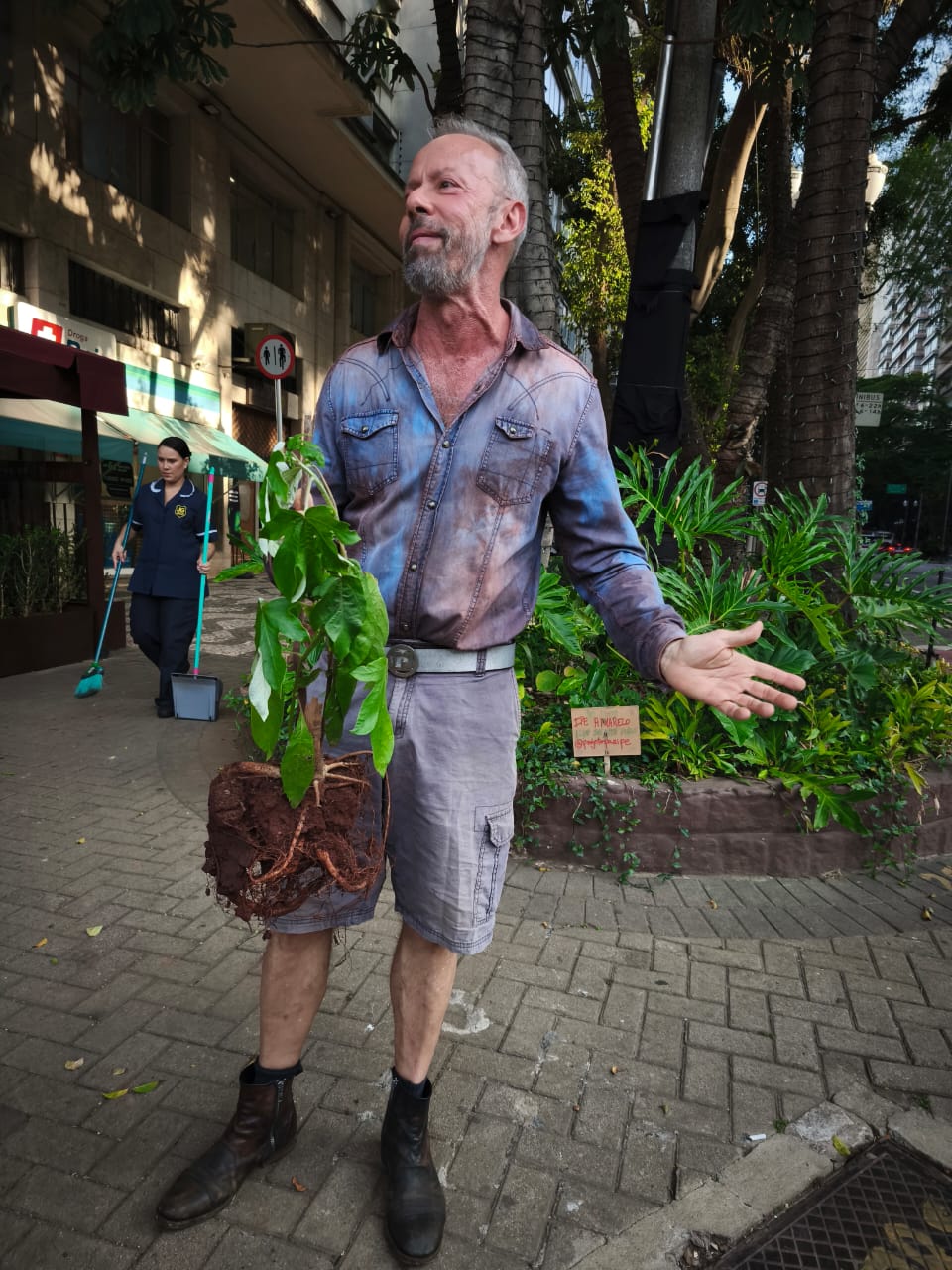 Imagem de Eduardo Pazian, o jardineiro da República, ele é um homem grisalho que veste uma camisa e shorts segurando uma muda de planta. 