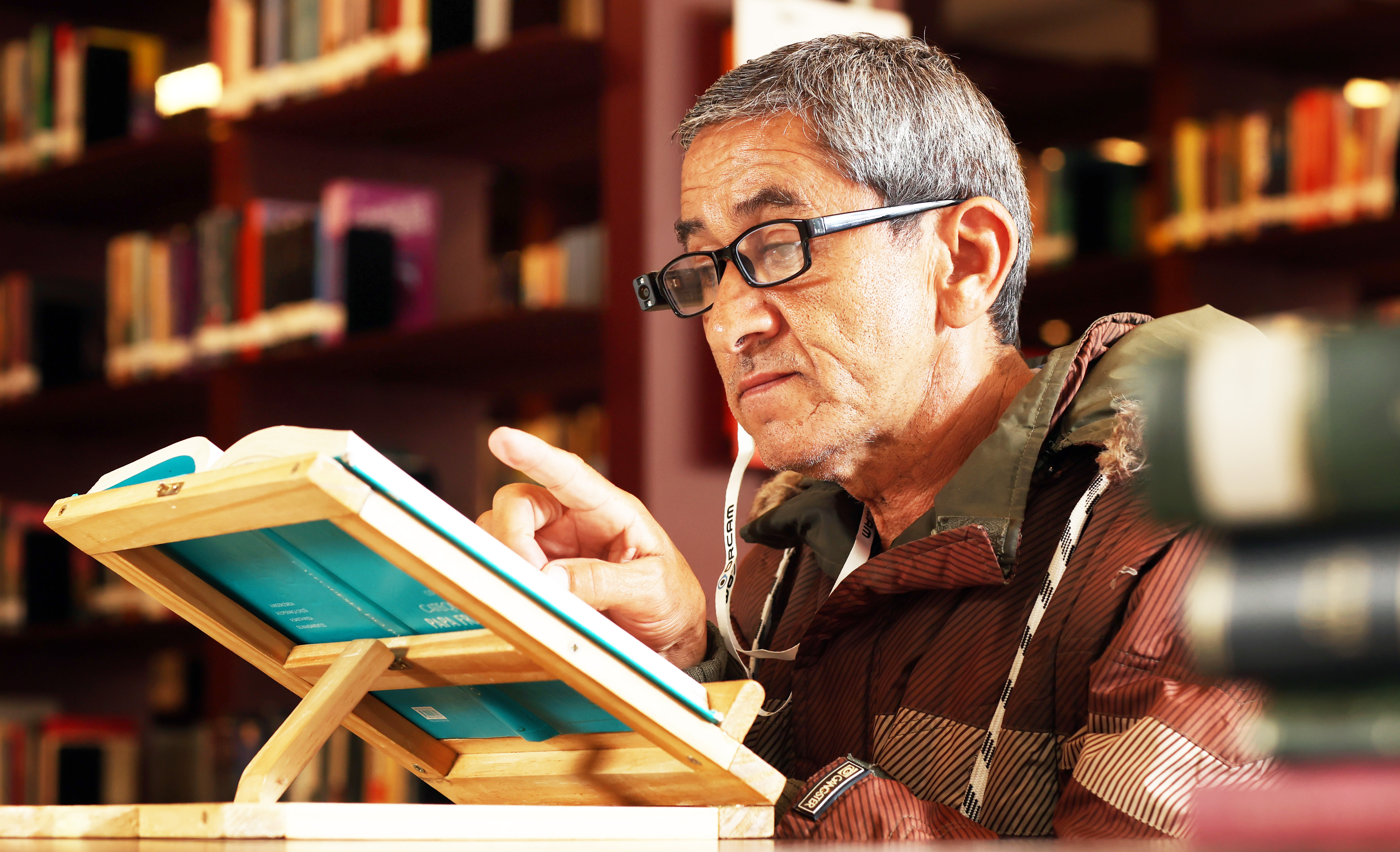 foto de um homem usando óculos inteligentes, ele é um homem idoso e está com uma expressão séria, com um livro apoiado em um suporte. 