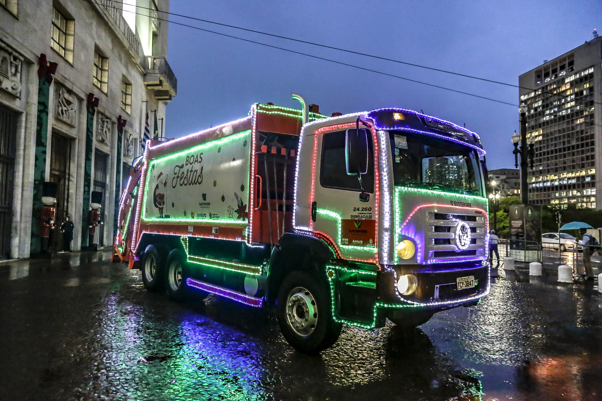 imagem de um caminhão de lixo decorado com luzes de Natal, ele está estacionado na frente da Prefeitura de São Paulo, o chão está molhada com água da chuva.