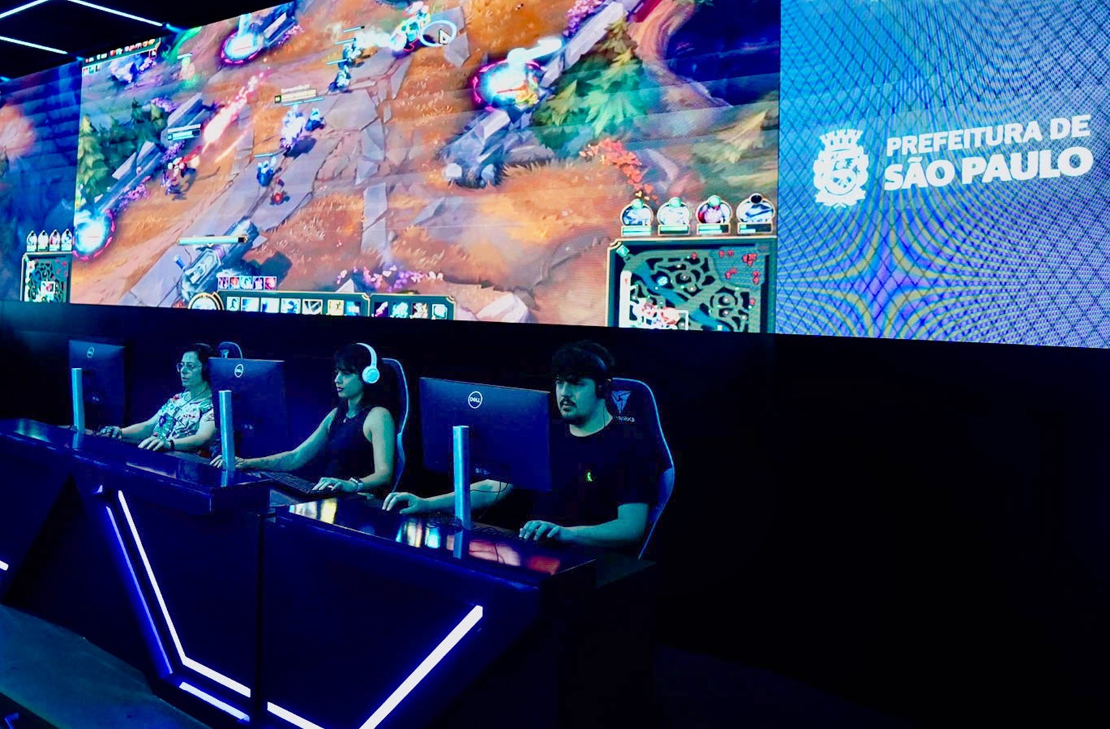 foto com uma grande bancada, onde 4 pessoas estão sentadas, elas usam headphone e atrás delas há um grande telão de led, com um fundo azul. 