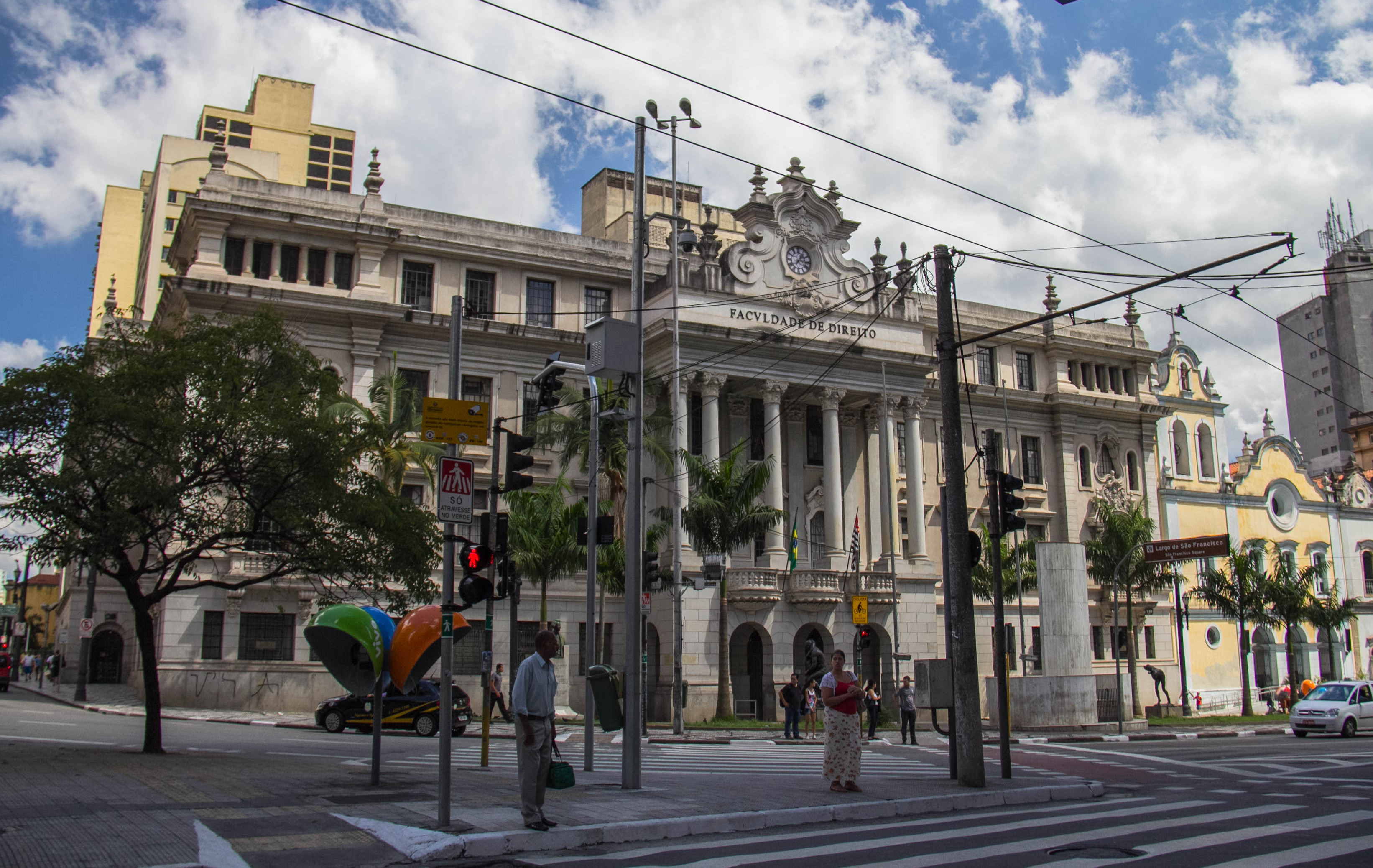 foto da entrada da Faculdade de São Francisco, um prédio no estilo clássico, com arcos na entrada, há um coqueiro na frente do prédio e pessoas andando. 