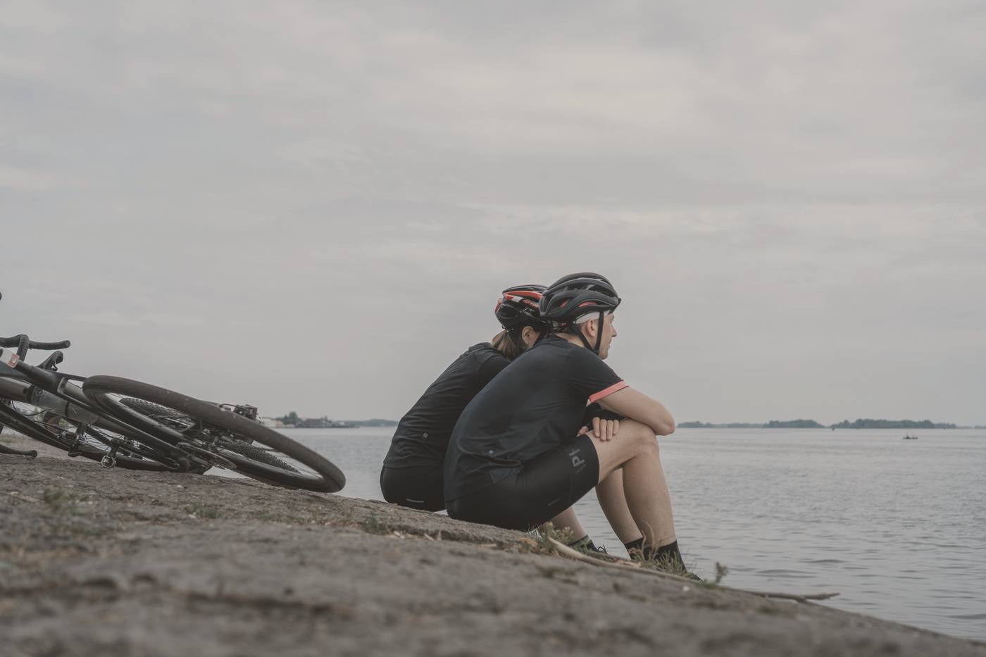 Two riders sitting by the water after a ride, bikes laid down beside them