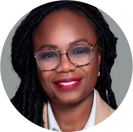Smiling woman with long black dreadlocks, wearing glasses, red lipstick, a beige blazer, and a white shirt against a gray background.