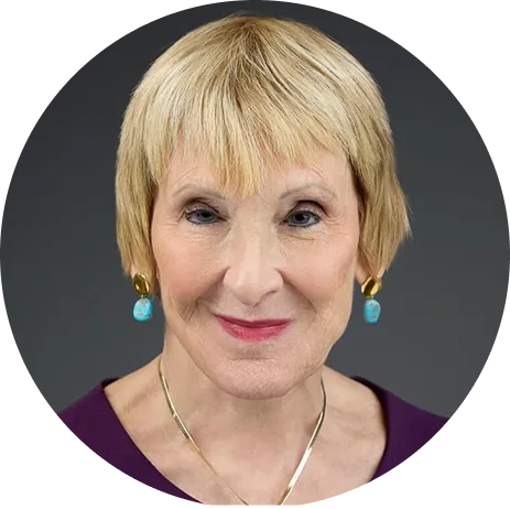 Portrait of an elderly woman with short blonde hair, wearing turquoise earrings and a purple top against a dark gray background.