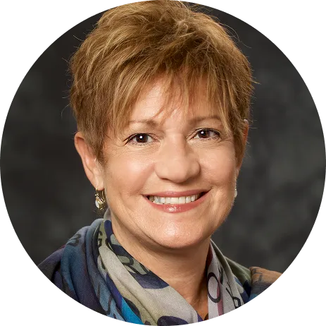 Smiling middle-aged woman with short light brown hair wearing a colorful scarf and earrings, against a dark blurred background.