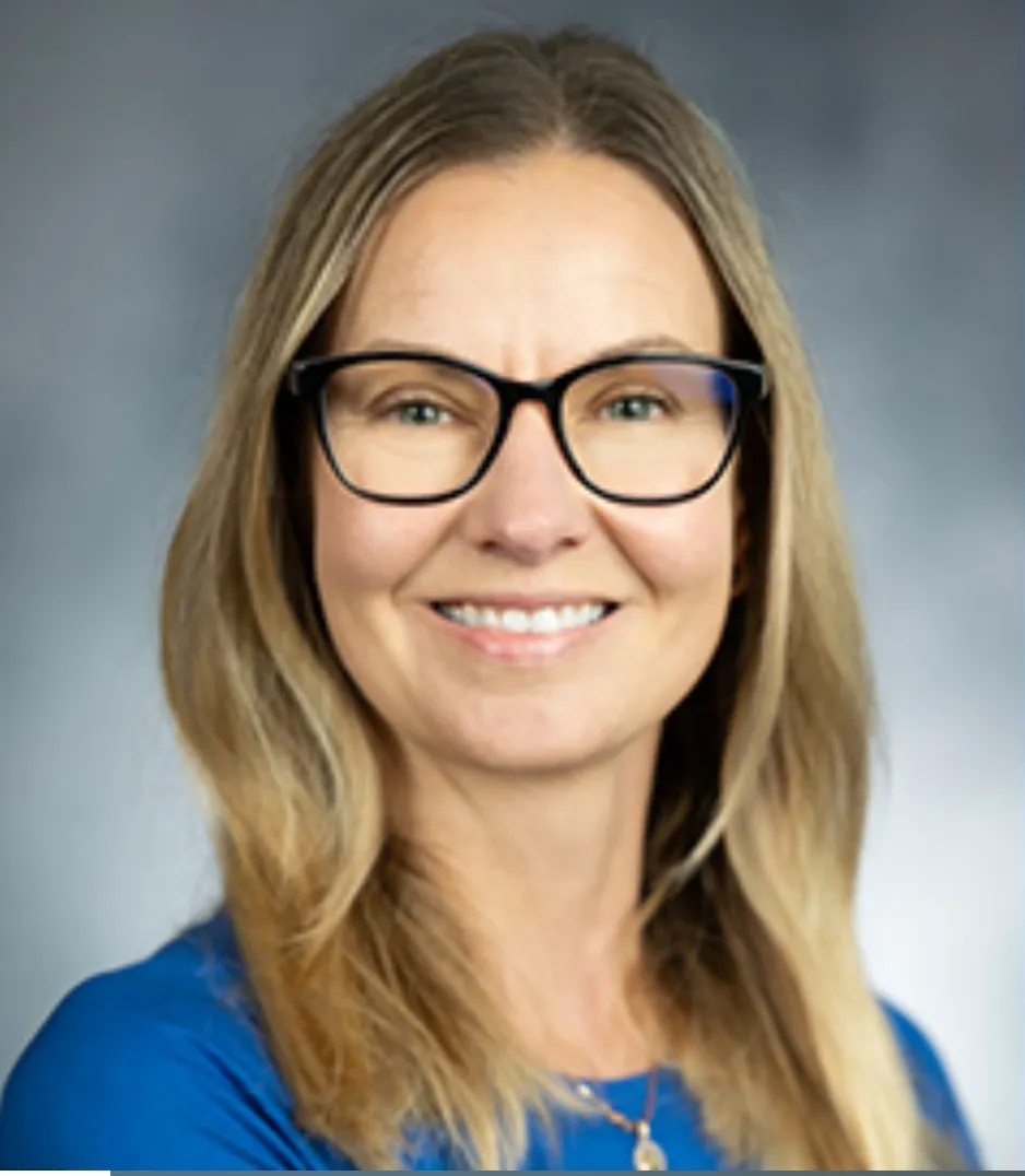 Smiling woman with long blonde hair wearing glasses and a blue top against a plain gray background.