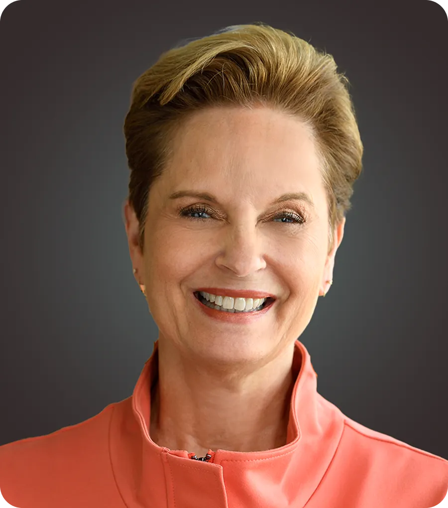 Smiling middle-aged woman with short blonde hair wearing a coral jacket against a dark gradient background.