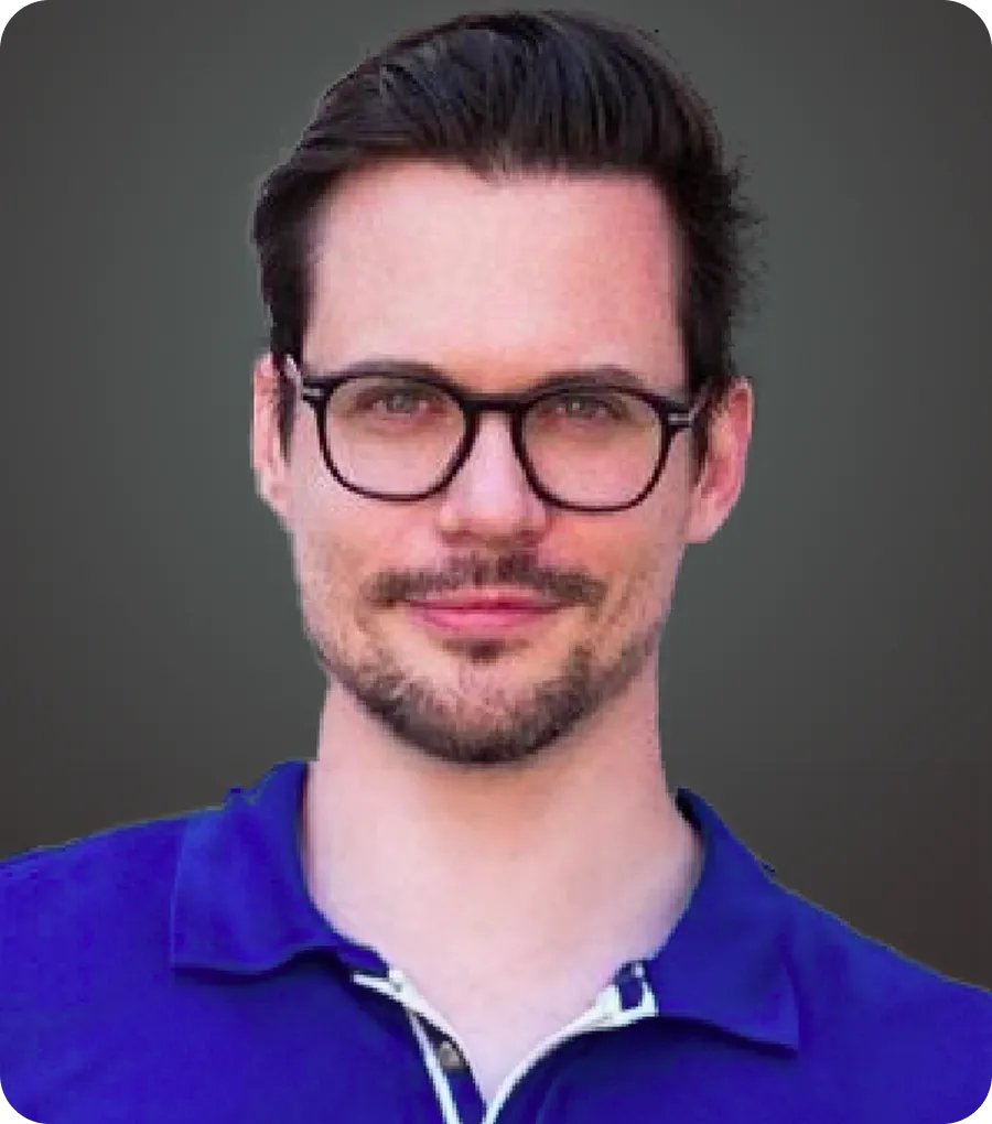 Man with dark hair and beard wearing black glasses and a blue collared shirt.