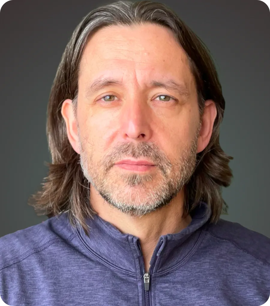 Middle-aged man with shoulder-length brown hair, light beard, and wearing a purple zip-up jacket, looking at the camera.