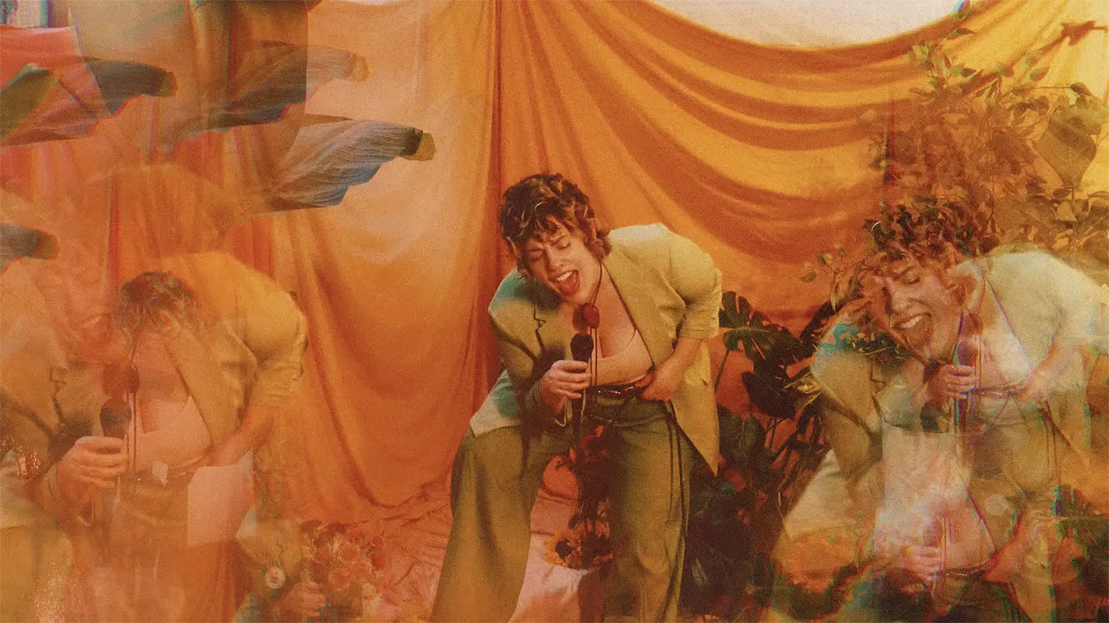 Warm, grainy video still of Jill in a green pantsuit standing in front of an orange drapery set with large green plants, leaning and singing into a mic