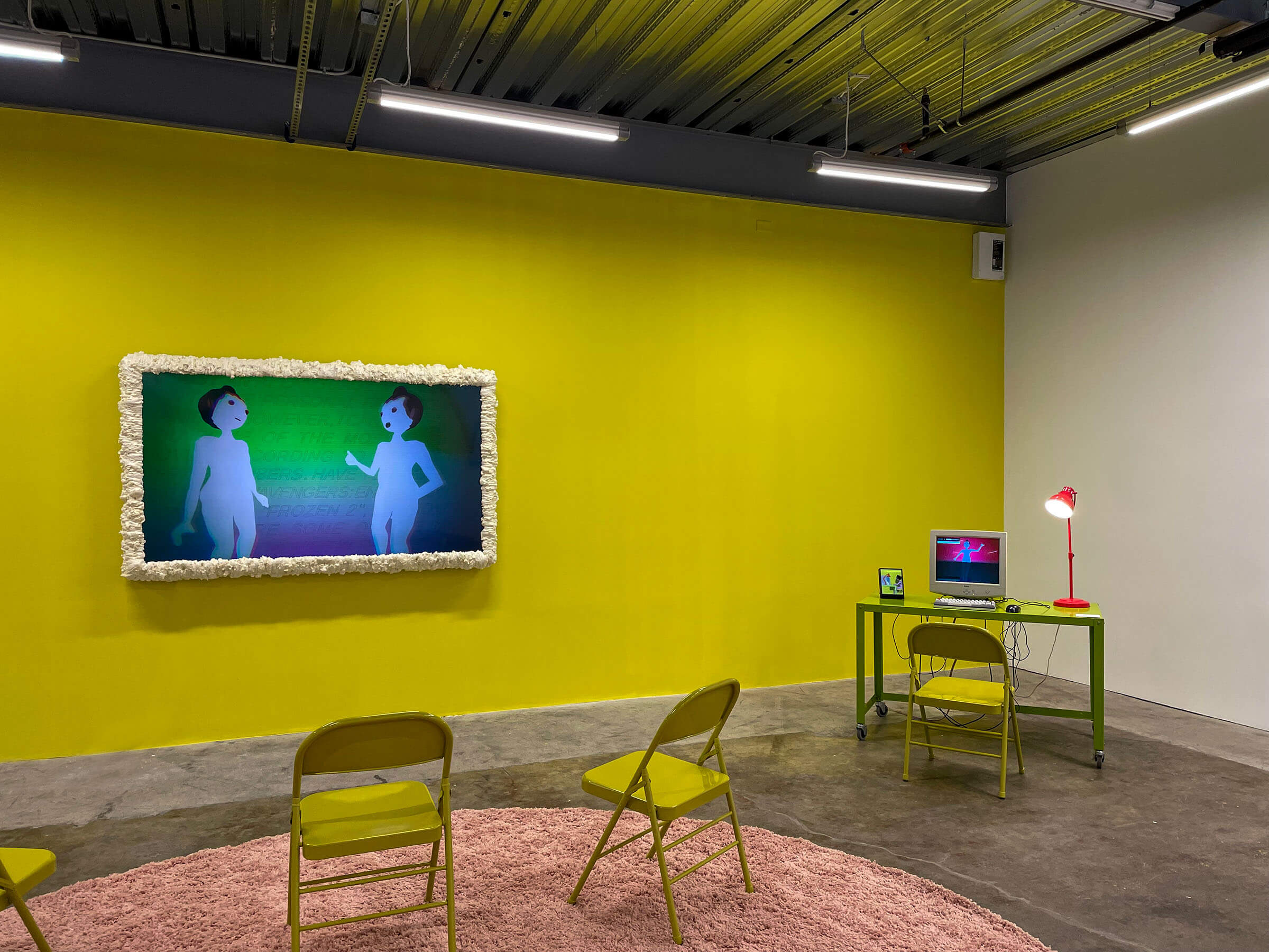 'SUPERPROMPT' installation view, showing a yellow wall with moulded TV screen playing an animation, with a green desk with computer also playing an animation