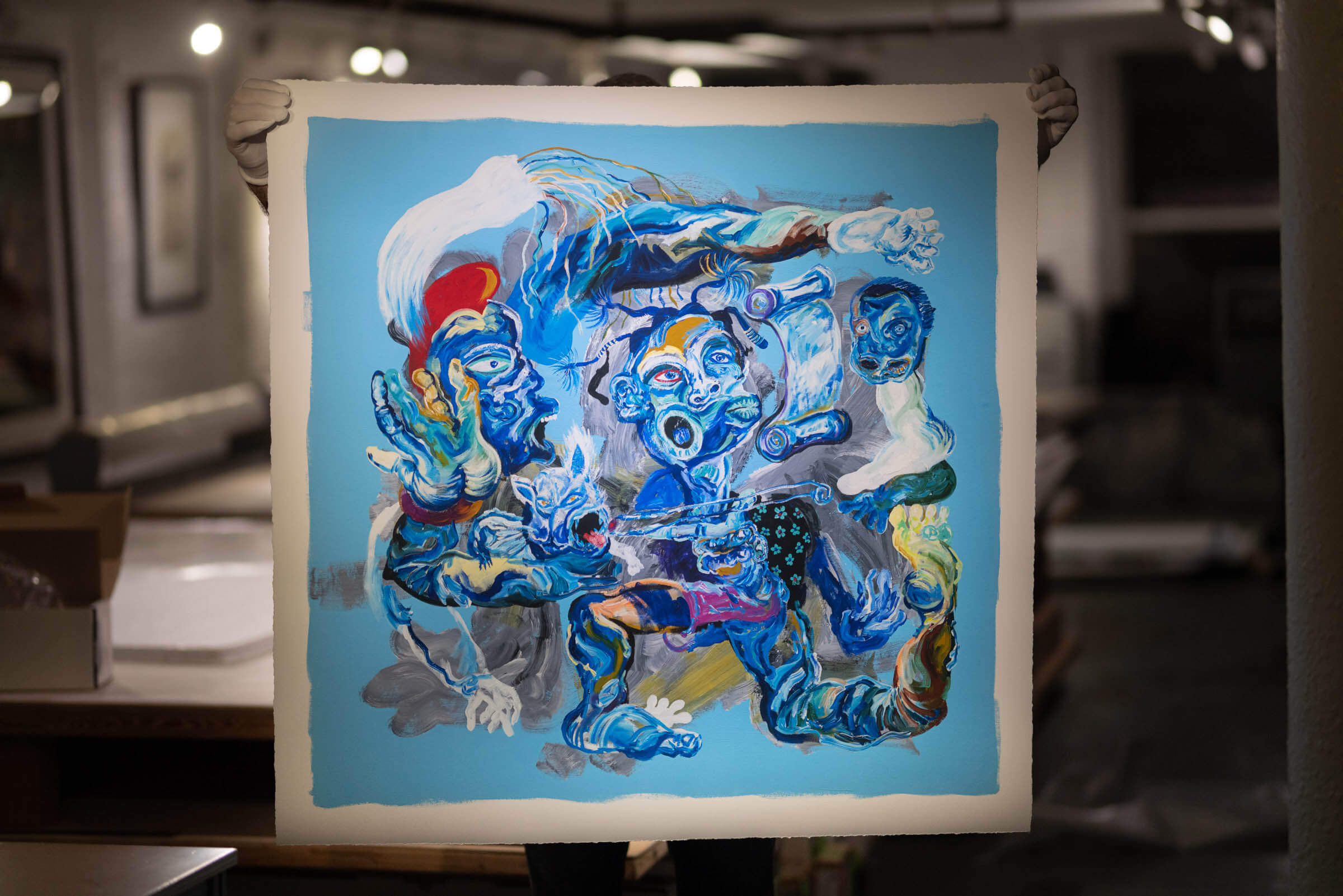 Holding up a print of Mangovo's Blue painting, featuring a disfigured human with 2 heads, morphing with animals and twisting its limbs to grasp scrolls