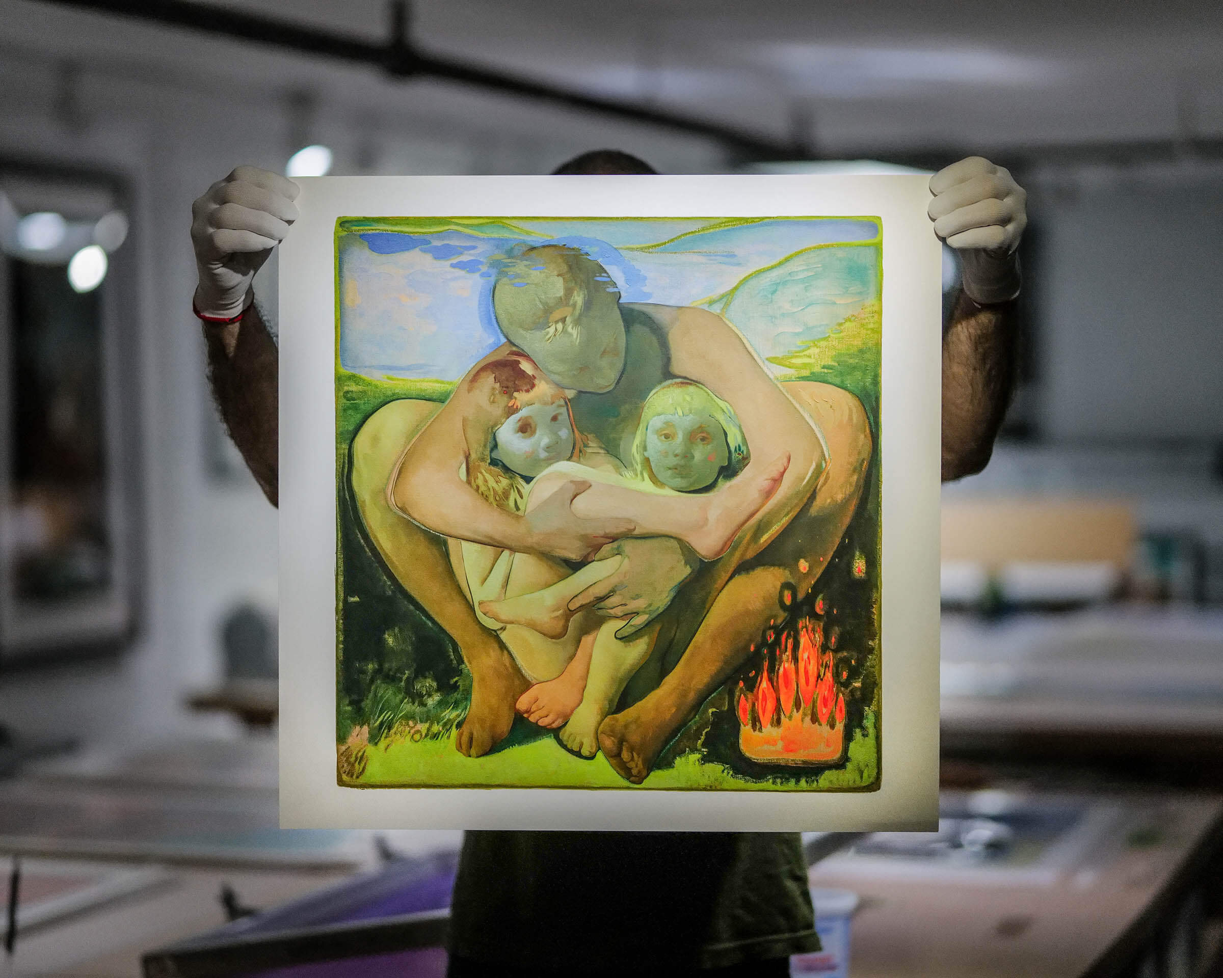 Holding up print 'Take This Fire' by Colleen Barry, that shows a maternal figure holding 2 infants next to a burning flame and set against a backdrop of hills and nature, painted in bright greens, yellows, and light blue colors