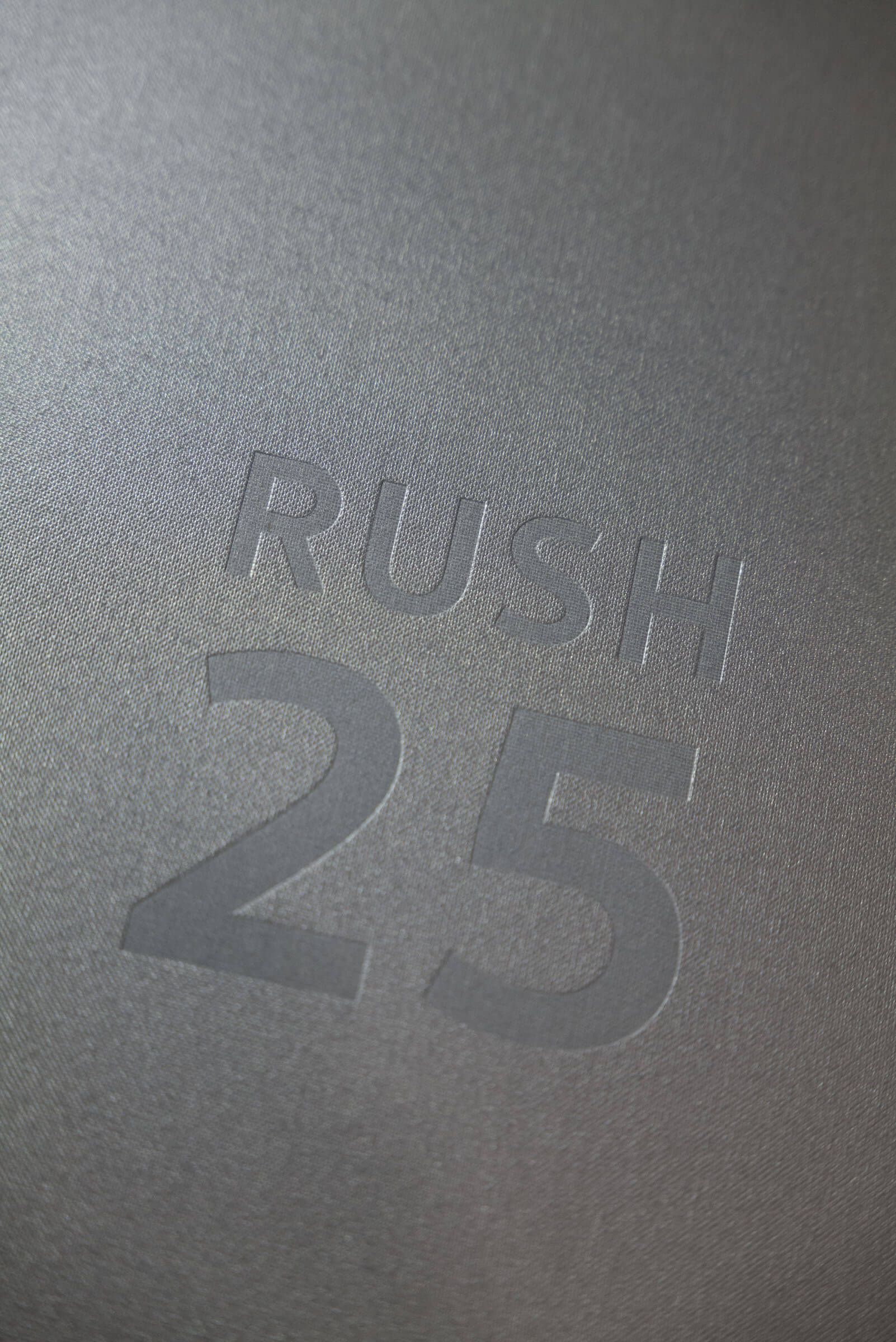Detail shot of the Rush Arts portfolio box cover, featuring a blind emboss that reads 'RUSH 25' on a coated silver fabric