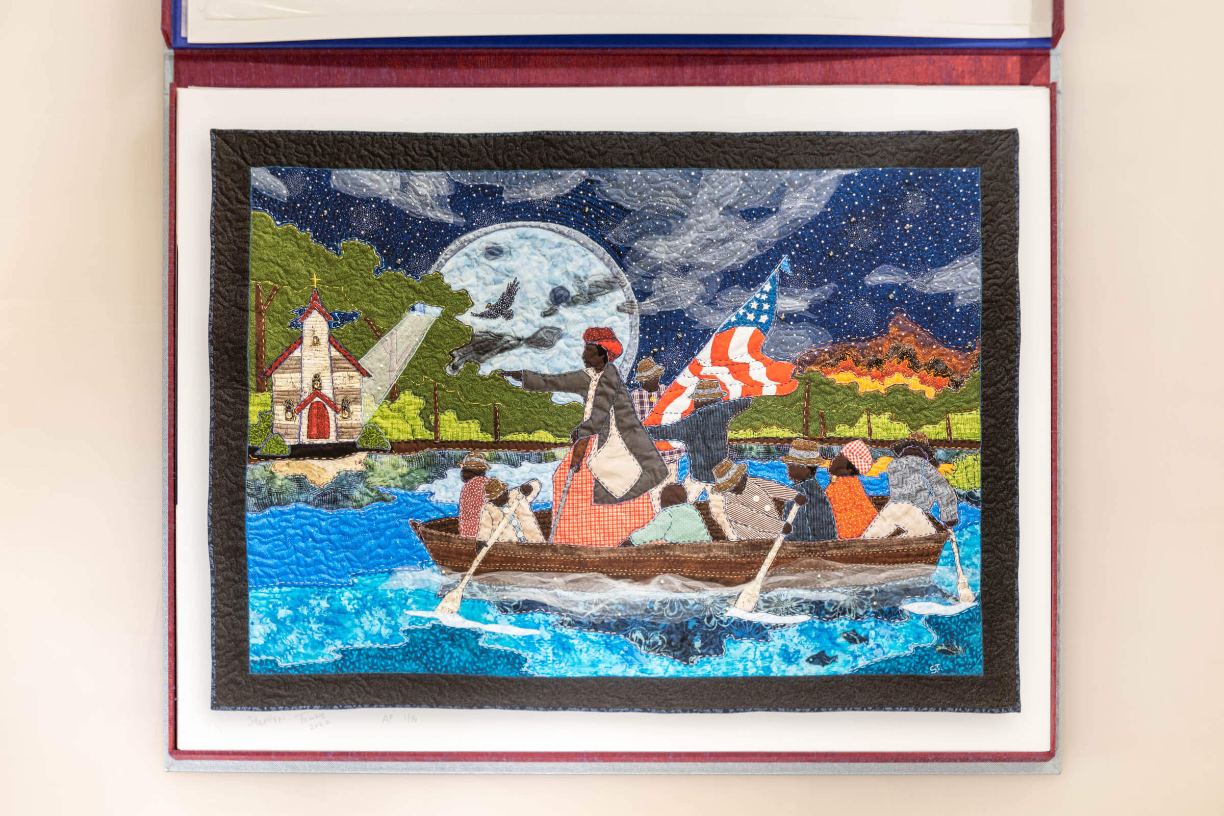 Portfolio box open to show Stephen Towns' 'We Shall Pass through The Combahee with You', which shows a stitched blanket of black figures carrying an American flag rowing a boat down a water body with hills, the sky, and buildings in the background