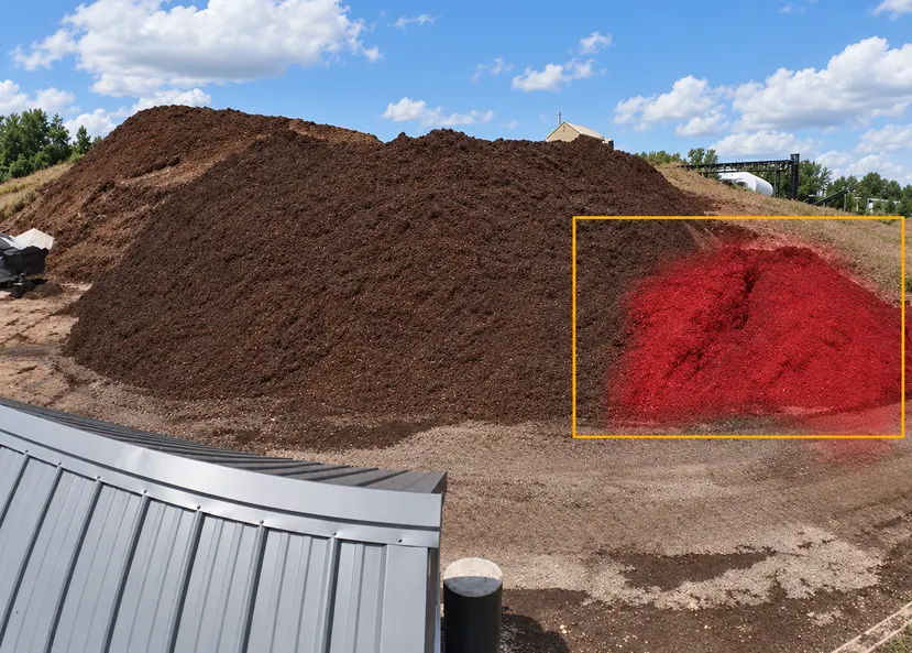 A large mound of dark compost fills an outdoor yard; a red-highlighted patch in a yellow box marks its right side, with a metal roof in front.