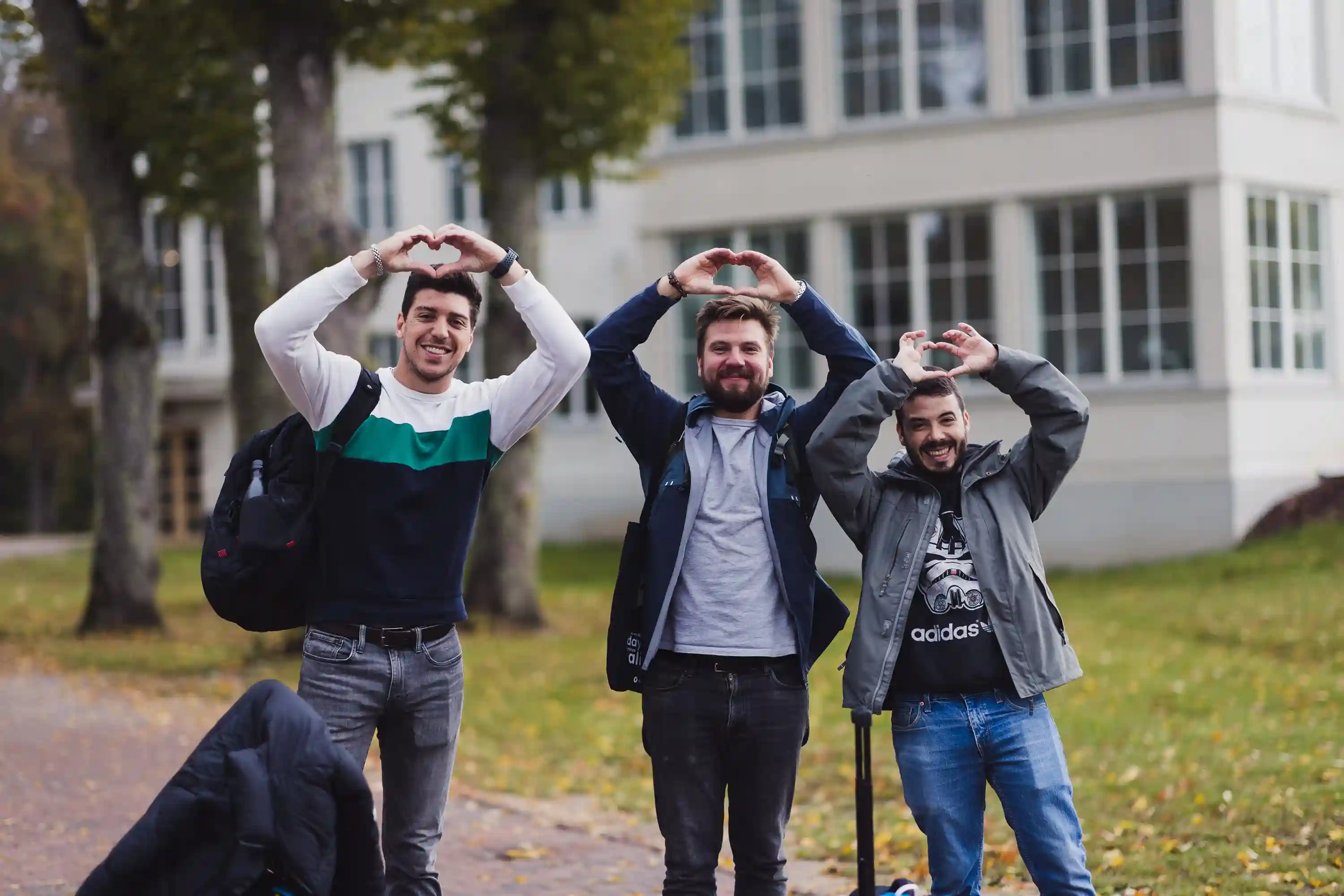 Three men outdoors form heart shapes with their arms, smiling widely. They stand on a leafy path beside a large building, exuding a cheerful and playful tone.