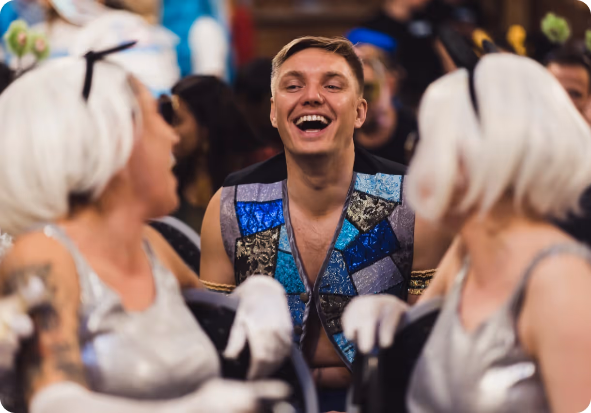 Smiling man in colorful vest laughing and engaging with two people in silver costumes and white wigs.