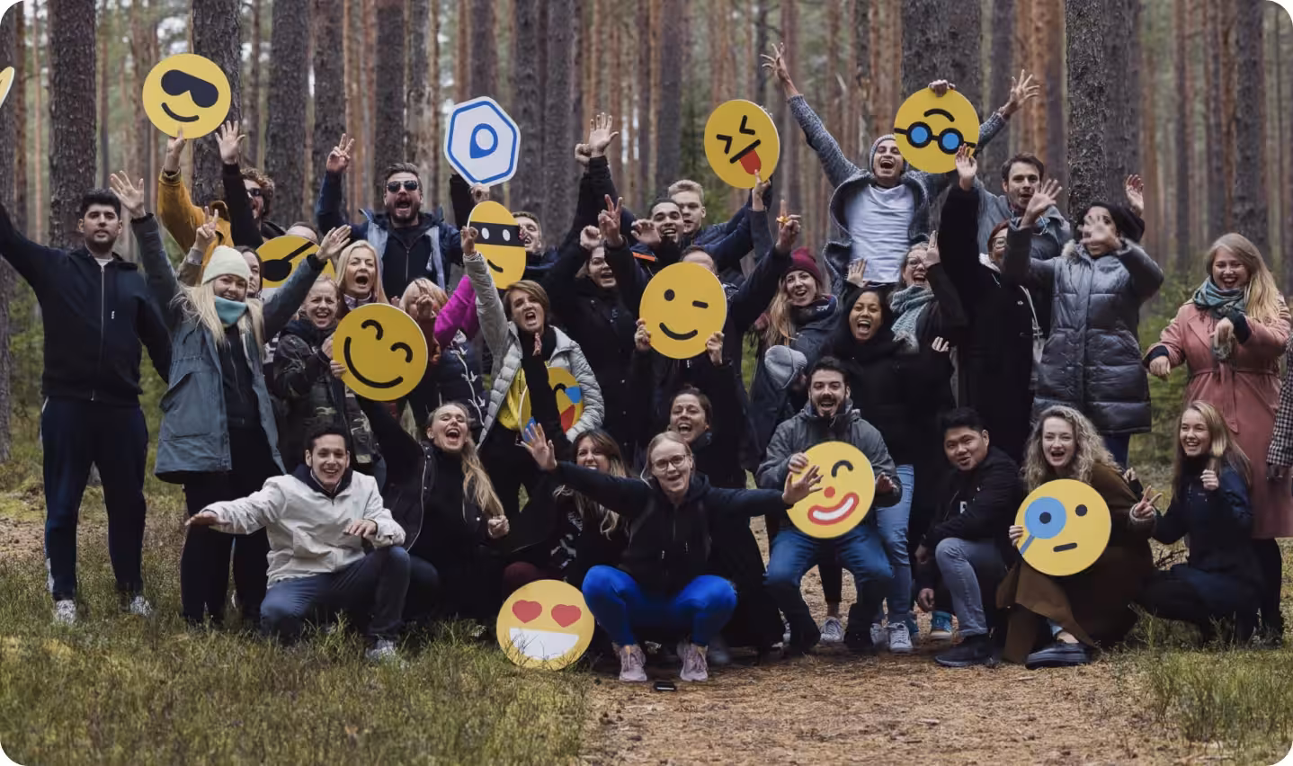 A joyful group of people in a forest, surrounded by tall trees, are enthusiastically posing with their arms raised. Emoji faces overlay their own, adding a playful and cheerful tone.