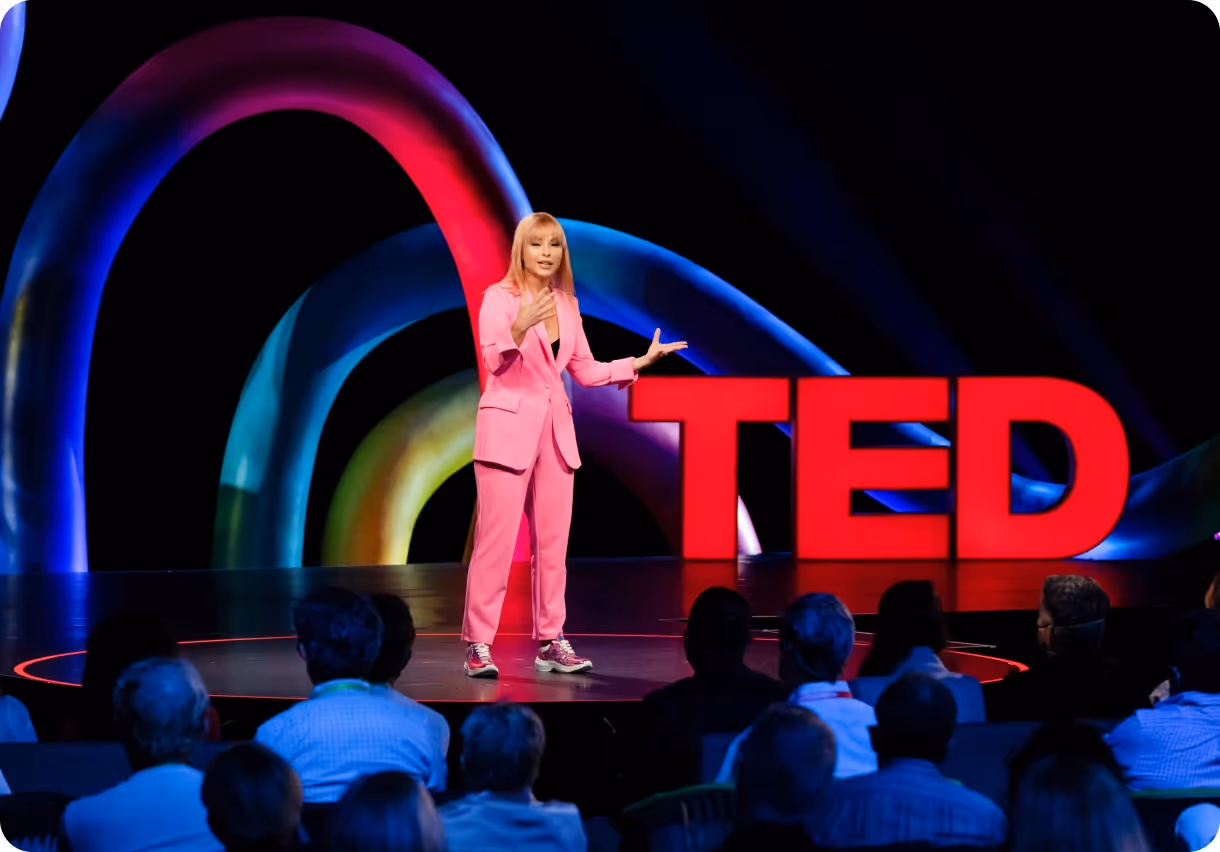 Speaker in a pink suit presenting on a stage with large red TED letters and colorful light arches in the background.