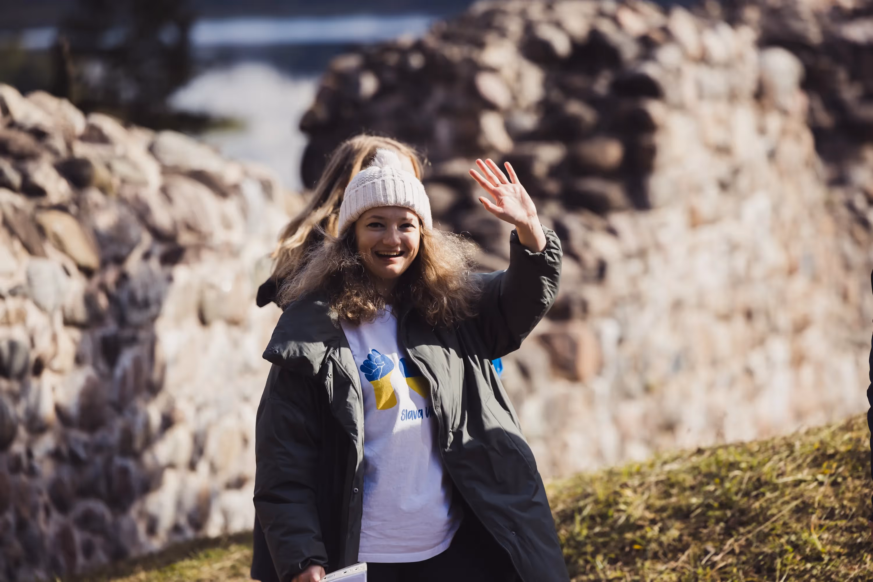 A woman in a winter coat and beanie smiles and waves, standing by a stone wall. Sunlight highlights her happy expression against the rustic backdrop.