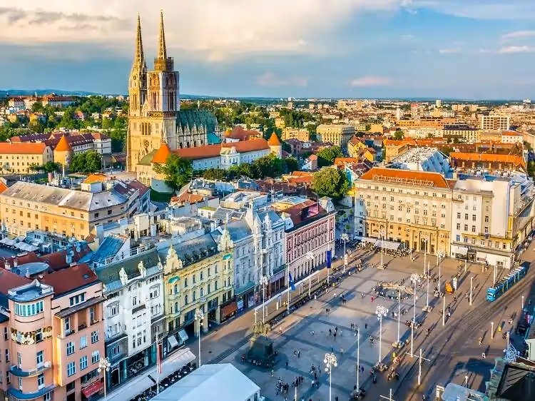 Aerial view of a vibrant cityscape featuring a historic cathedral with twin spires, surrounded by colorful buildings and a bustling plaza under a clear sky.