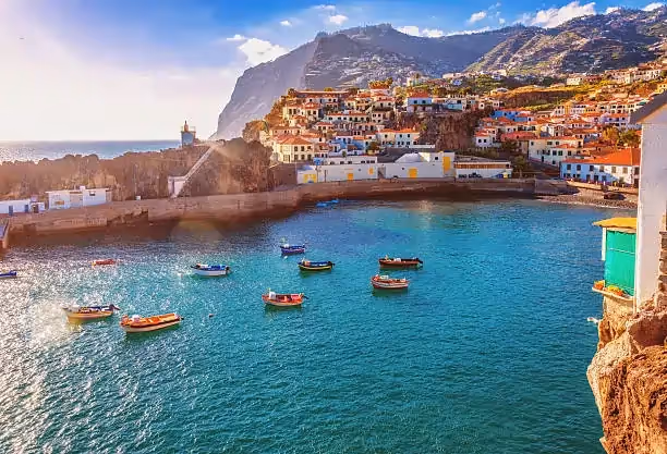 Scenic coastal village with colorful boats floating on turquoise water, nestled against rocky cliffs with sunlit houses and distant mountains. Tranquil and picturesque.